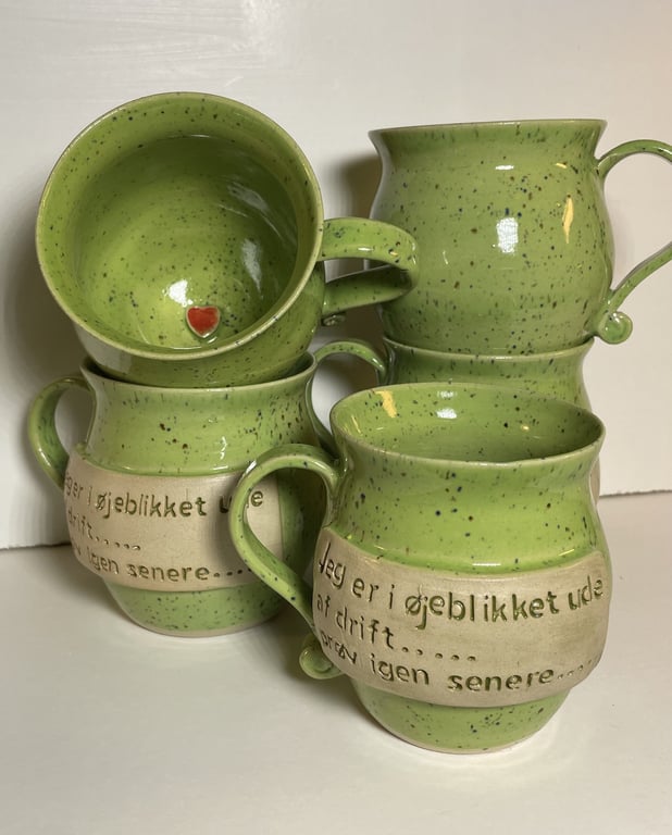 Fire grønne keramik kopper med beige bånd og tekst på dansk, stablet mod en hvid baggrund. En kop viser en lille rød hjerteformet detalje indeni.