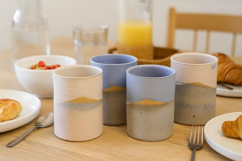 Fire keramikkrus med blå og beige design på et morgenbord med croissanter, en bolle med yoghurt og frugt, appelsinjuice og bestik.