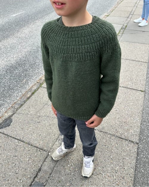Dreng iført en grøn strikket sweater, mørke bukser og hvide sneakers, stående på en fortov ved en vej. (2)
