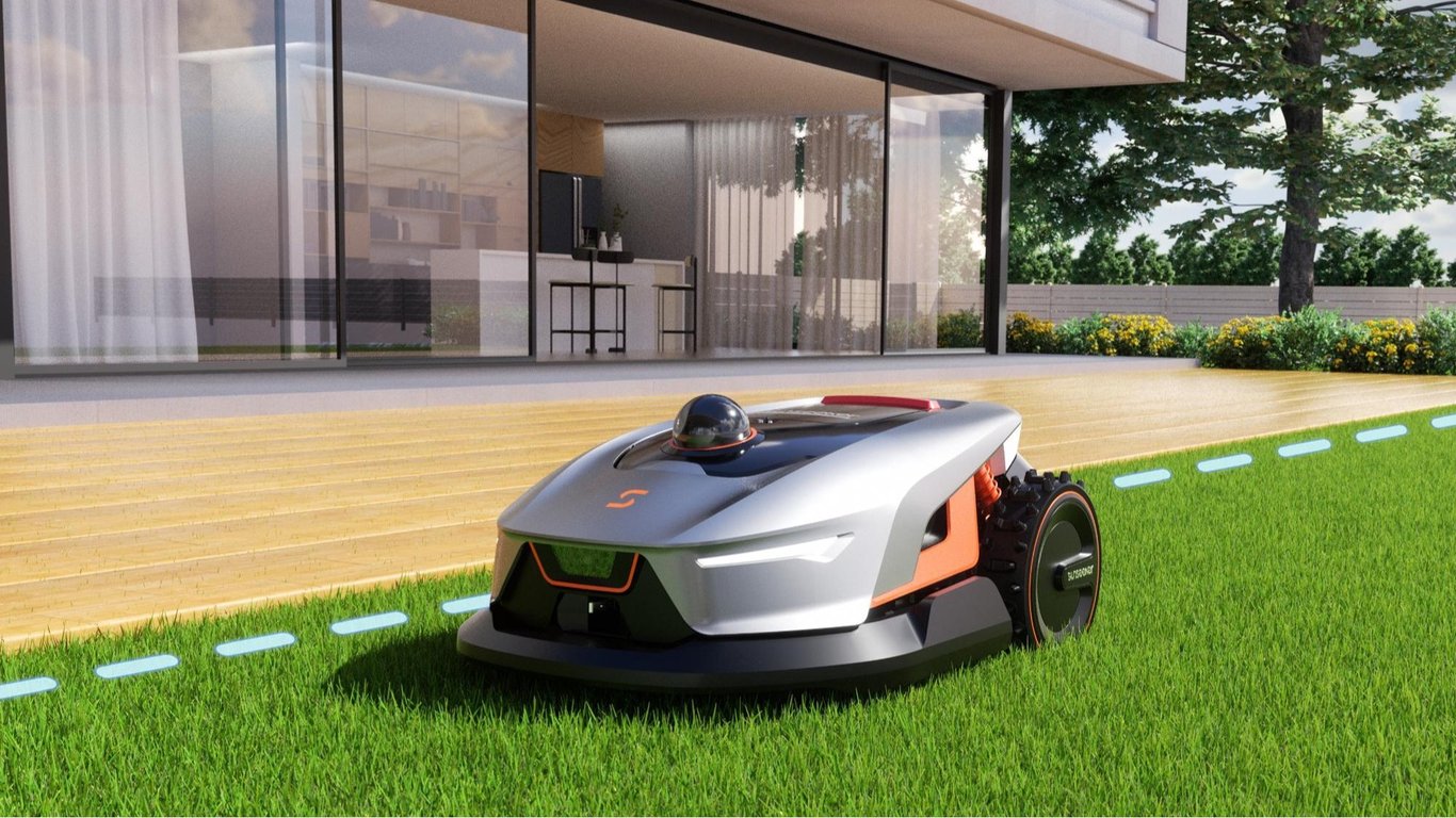 Robotplæneklipper, der kører på græsplæne foran moderne hus med glasfacade og træterrasse.