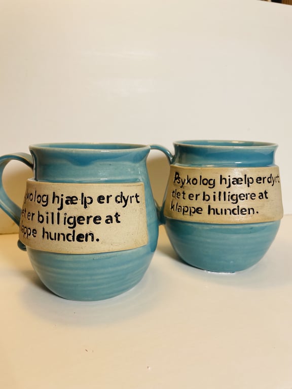 To blå keramik krus med hank med teksten "Psykologhjælp er dyrt, det er billigere at klappe hunden." skrevet på en beige plade på forsiden af hvert krus.