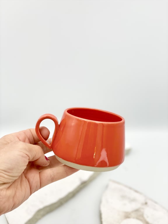 Hånd holder en rød keramikkop med hank mod en lys baggrund.