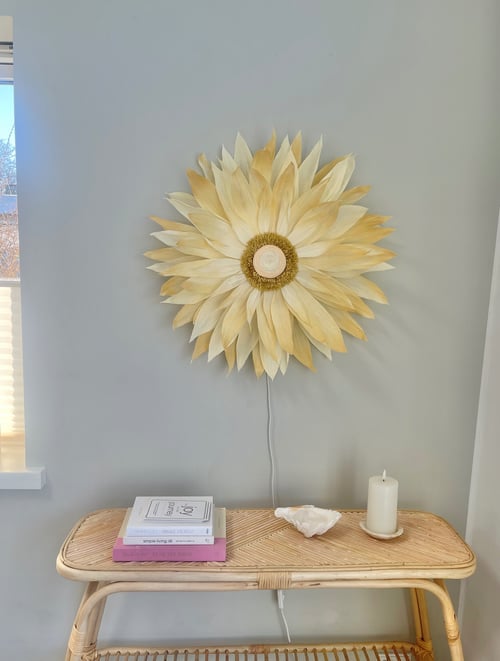 Væglampe formet som en stor, lys solblomst over et lyst flettet bord med bøger, en konkylie og et hvidt stearinlys på en lys baggrund. (1)