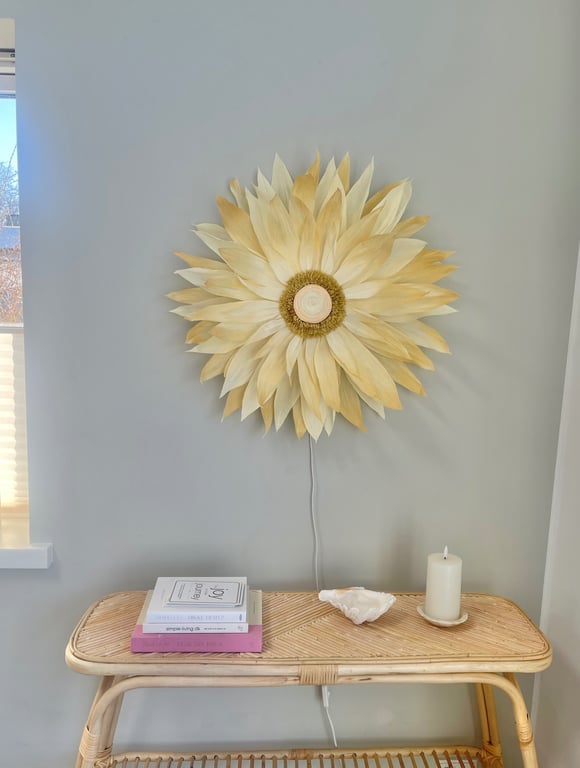 Væglampe formet som en stor, lys solblomst over et lyst flettet bord med bøger, en konkylie og et hvidt stearinlys på en lys baggrund.