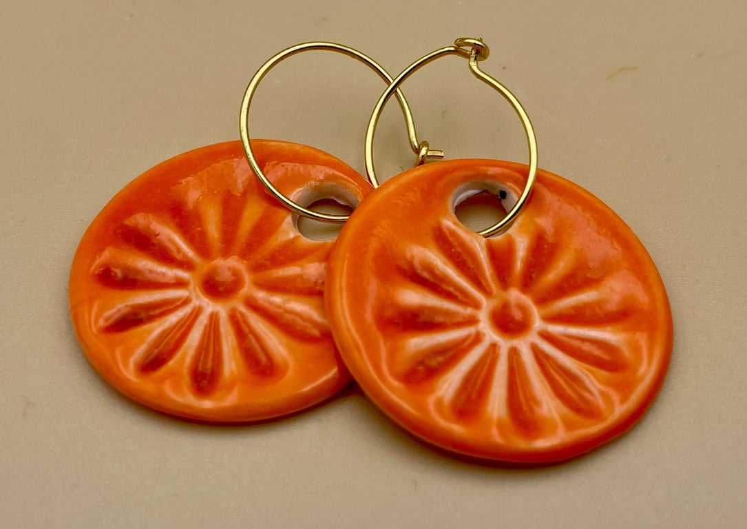 To orange keramikøreringe med blomsterrelief og guldbøjler liggende på en beige overflade.