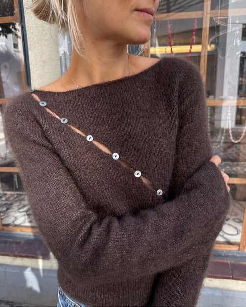 Kvinde med lyst hår iført brun, strikket sweater med skrå række af knapper og slidser, står foran et vindue. (3)