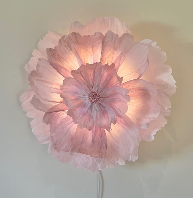 Blomstformet væglampe med bløde, lyserøde og hvide kronblade, der lyser varmt op mod en lys væg.