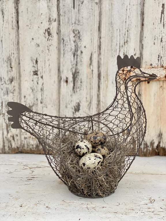 Trådkurv formet som en høne med fire pyntede æg og dekorativ mose indeni, placeret foran en slidt trævæg.