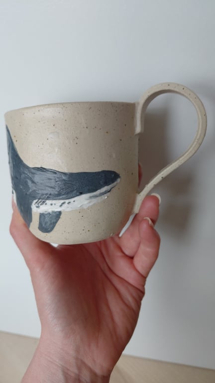 Hånd, der holder en beige keramikkop med et malet motiv af en hval.