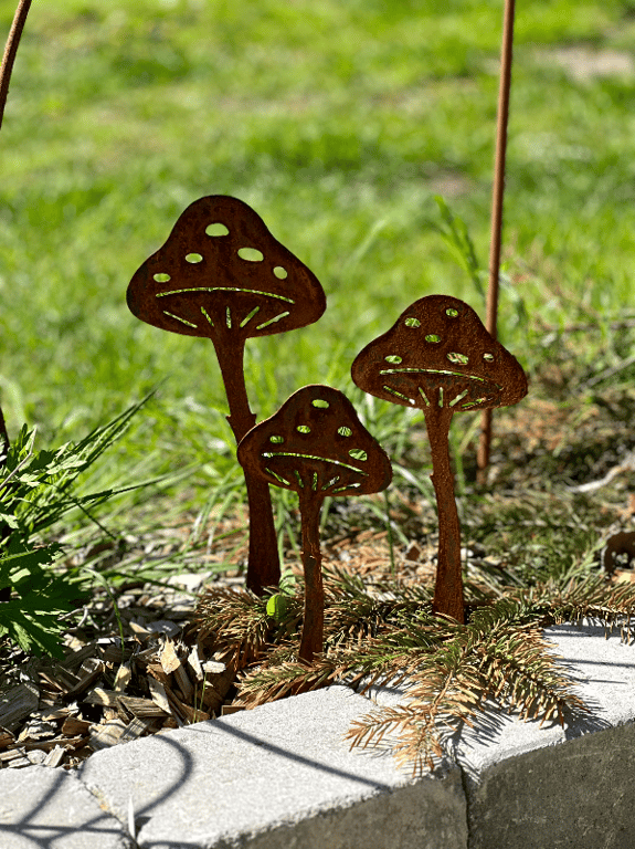 Rustfarvede metaldekorationer formet som tre svampe i en have omkranset af græs og sten.