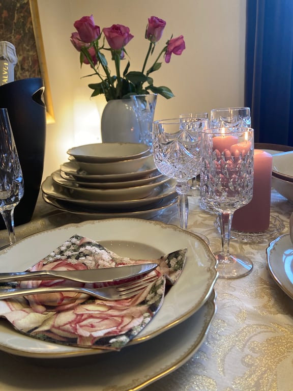 Dækket spisebord med fine porcellens tallerkener, bestik på blomsterprintet serviet, krystalglas, levende lys og en vase med lyserøde roser. (2)
