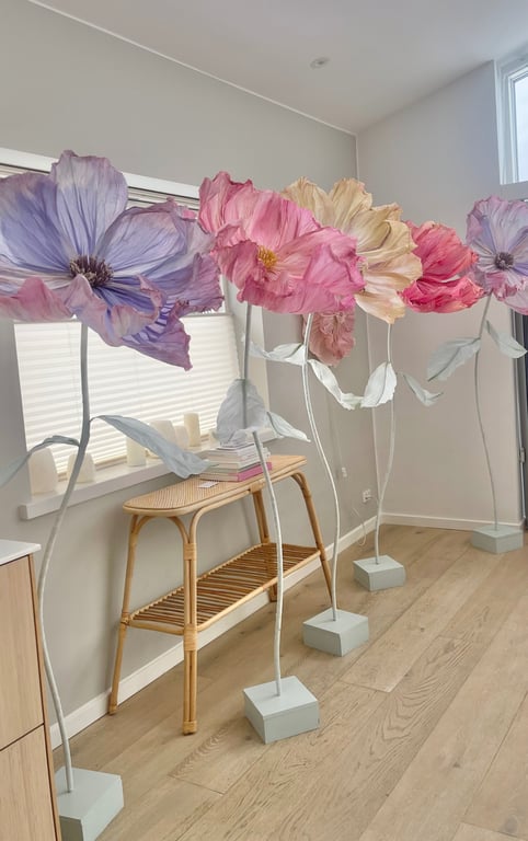 Fem store kunstige blomster i pastelfarver står på rækker i et lyst rum med trægulv og hvide vægge.
