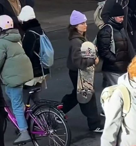 En gruppe mennesker iført vintertøj, hvor en kvinde med en lilla hue bærer et skateboard under armen, mens en person i en grøn jakke sidder på en cykel. (1)