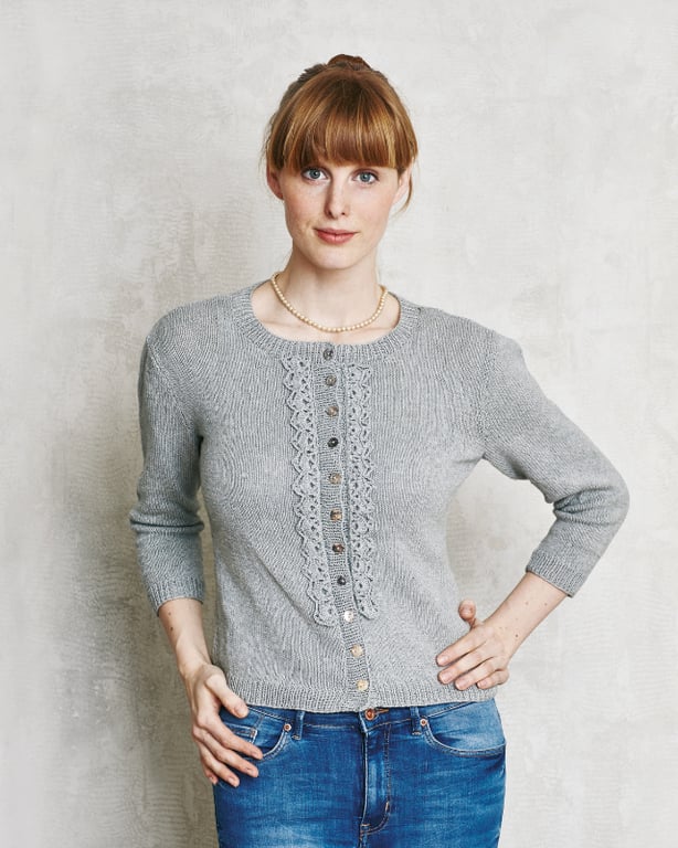 Kvinde iført grå strikket cardigan med knapper og perlehalskæde, poserende foran beige væg.