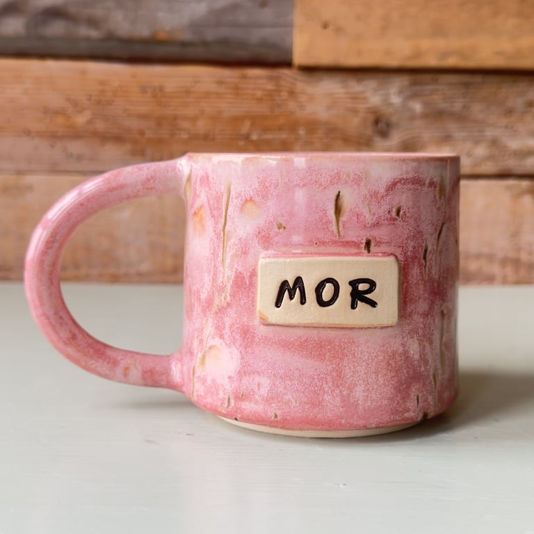 Rosa keramisk kop med teksten "MOR" på en beige plade foran, stående på et lyst bord med en træbaggrund.