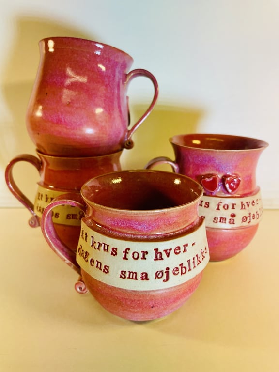 Billede af fire pink keramikkrus med hank, hvorpå der står tekst på dansk i hvidt bånd rundt om midten.
