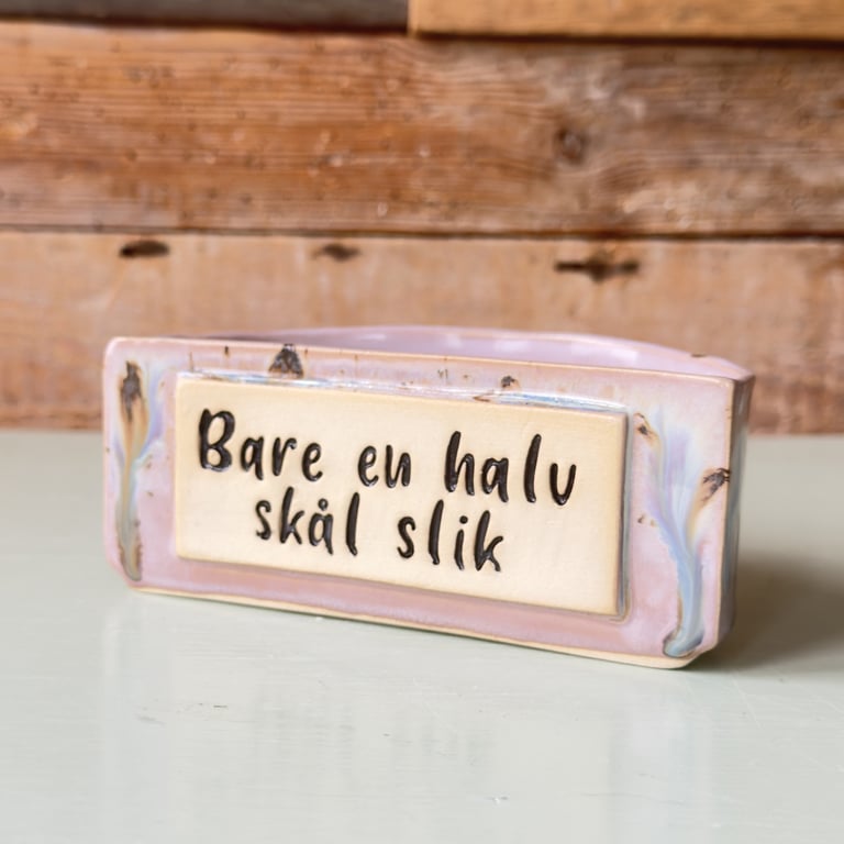 Keramisk skål med teksten "Bare en halv skål slik" på en rektangulær plade, placeret på en hvid bordplade med en trævæg i baggrunden.