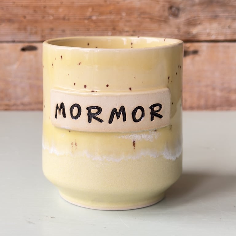 Gul keramikkop med teksten "MORMOR" i sort på en beige rektangulær plade, placeret på en hvid overflade med en træbaggrund.