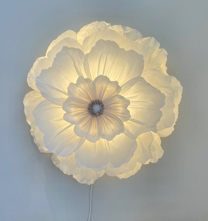 Væglampe formet som et stort, hvidt blomstermotiv med bløde, lagdelte kronblade, der lyser varmt.