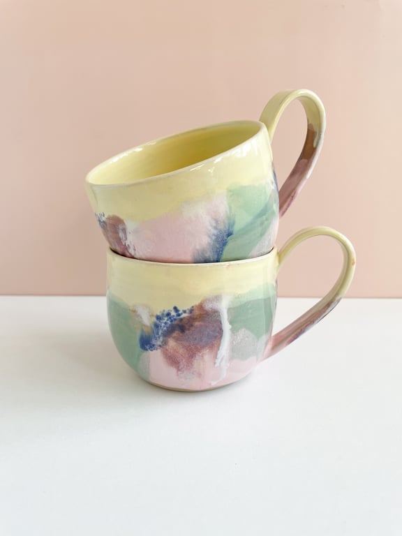 To malede keramikkopper i pastelfarver med abstrakte mønstre stablet ovenpå hinanden mod en rosa baggrund.