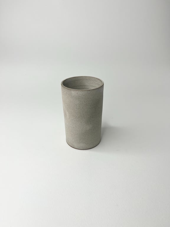 Grå, cylindrisk keramisk kop stående på en hvid overflade med ensfarvet baggrund.