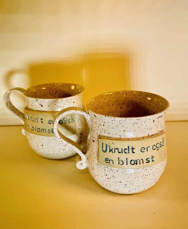 To kopper med prikmønster og teksten "Ukrudt er også en blomst" på en lys overflade.