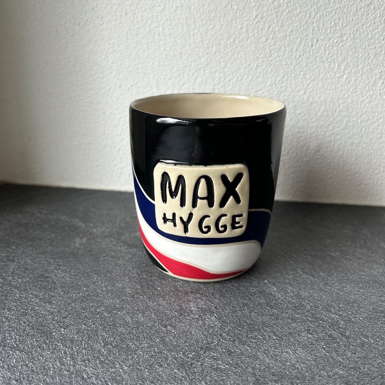 Sort keramik kop med teksten "MAX HYGGE" og farvede bølgemønstre i blå, hvid og rød, placeret på gråt bord mod hvid væg.