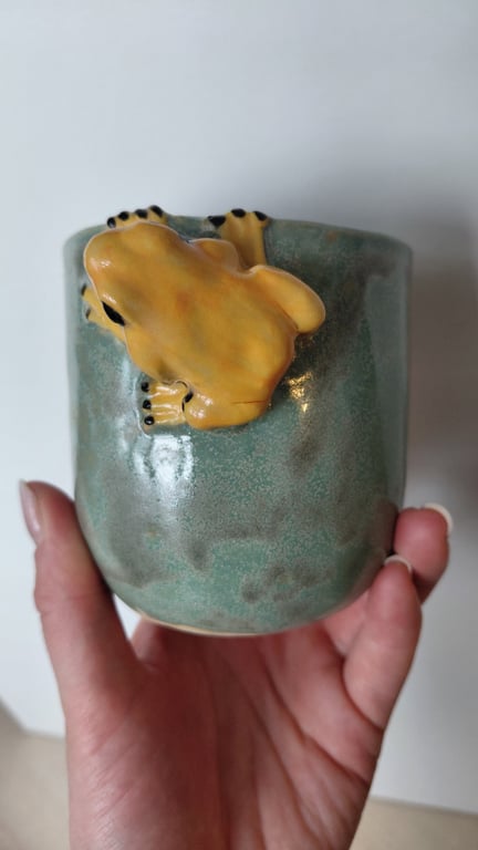 Hånd holder en grøn keramik kop med en gul keramisk frø, der klamrer sig til koppen.