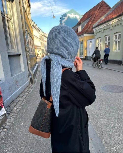 Person iført sort jakke og lyseblåt strikket tørklæde, gående på gade med traditionelle bygninger og cyklister i baggrunden. (5)
