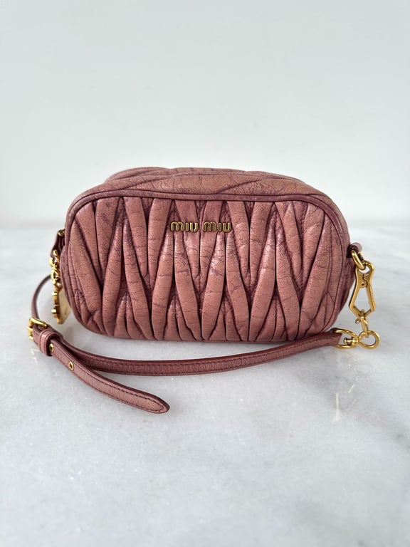 Lille pudderfarvet Miu Miu taske med quiltet læder og guldfarvede metalbeslag, placeret på hvid marmoroverflade.