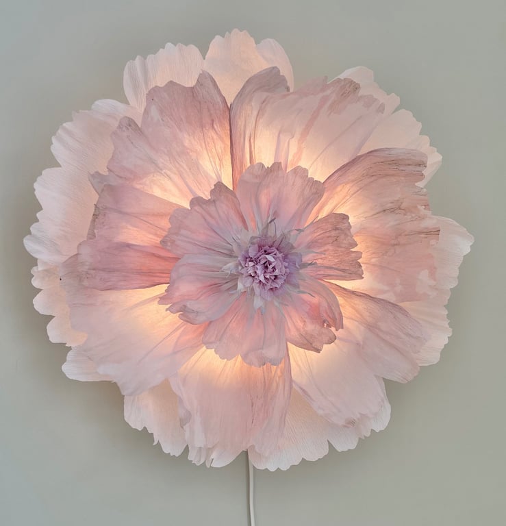 Blomstformet lampe med bløde lyserøde blomsterblade oplyst indefra på en grå væg.