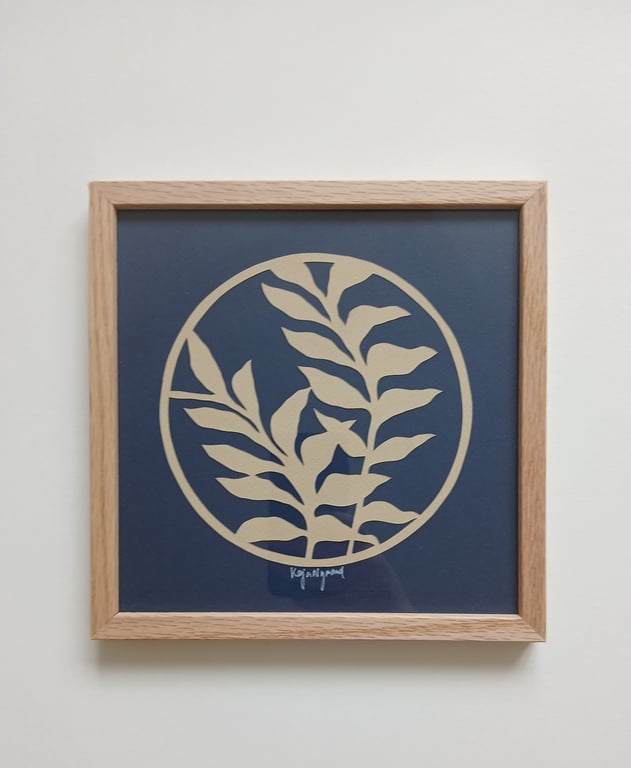 Tavlebillede med en simpel, beige plante med blade indrammet i lyst træ mod en mørkeblå baggrund.