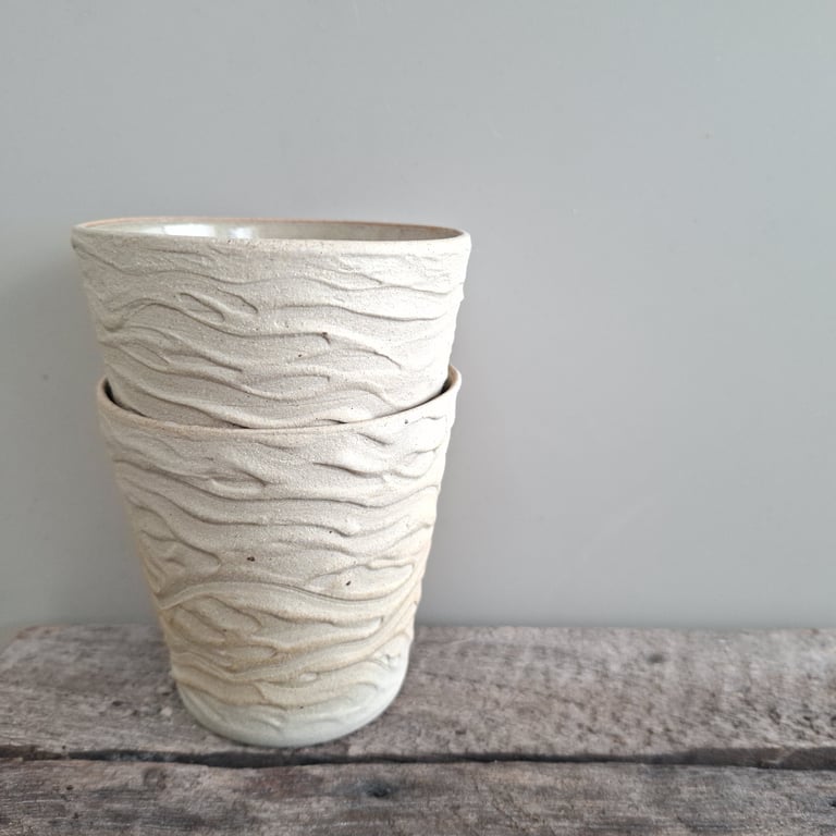 To kopper i keramik med bølget tekstur, stablet oven på hinanden på en grov træflade foran en grå væg.