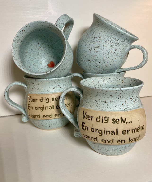 Billede af fem lyseblå keramikkrus med brunlige pletter og en beige tekststrimmel med dansk tekst "Vær dig selv... En original er mere værd end en kopi".