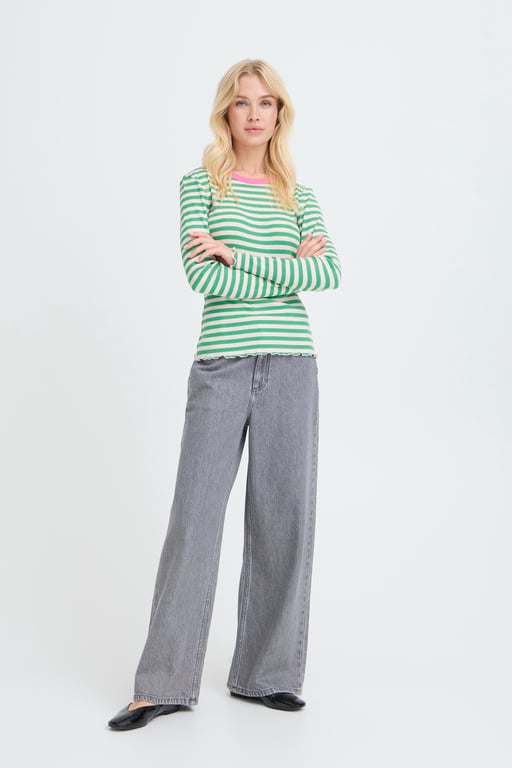 Kvinde iført grøn- og hvidstribet langærmet bluse med lyserød kant, grå vide bukser og sorte flade sko, stående med armene krydset.