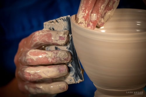 Nærbillede af hænder dækket af ler, der former en drejet lerkrukke med en skraber.