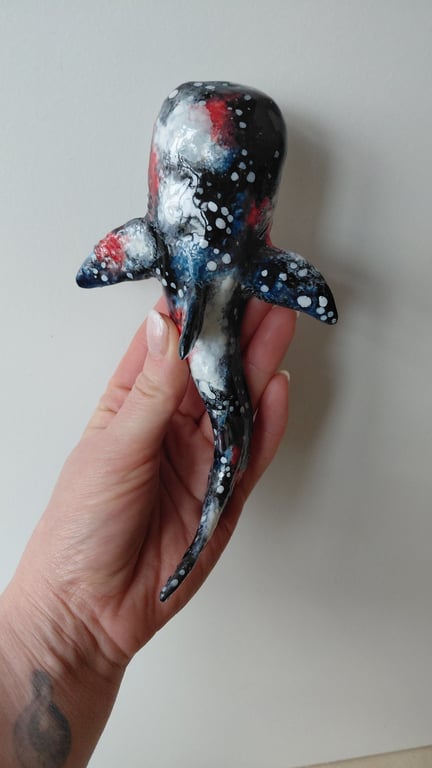 Hånd holder en lille, håndmalet skulptur af en hvalhaj med sort, rød, blå og hvid farvetryk, på lysegrå baggrund.