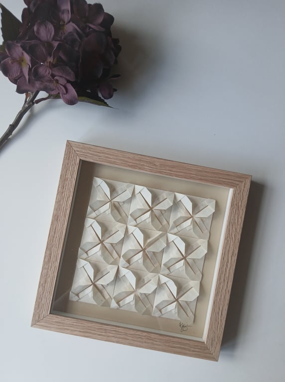 Billedramme med origamiblomster i beigefarvet papir, placeret på hvid overflade ved siden af en mørklilla tørret blomst.