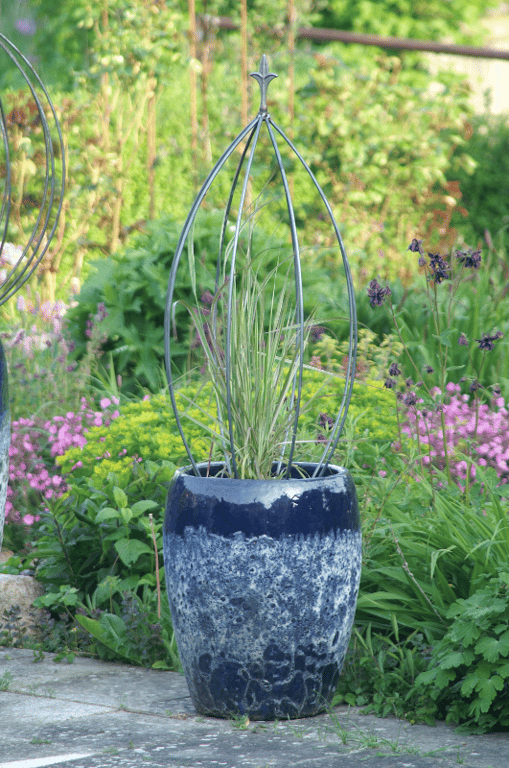 En stor blå keramisk urtepotte med et metalbur og grønt græsplante, placeret på en stenbelagt havegang med blomstrende planter og grønne buske i baggrunden.