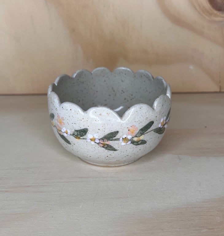 Keramisk skål med bølget kant og blomsterdekor med grønne blade og små hvide og orange blomster, placeret på et lyst træbord.