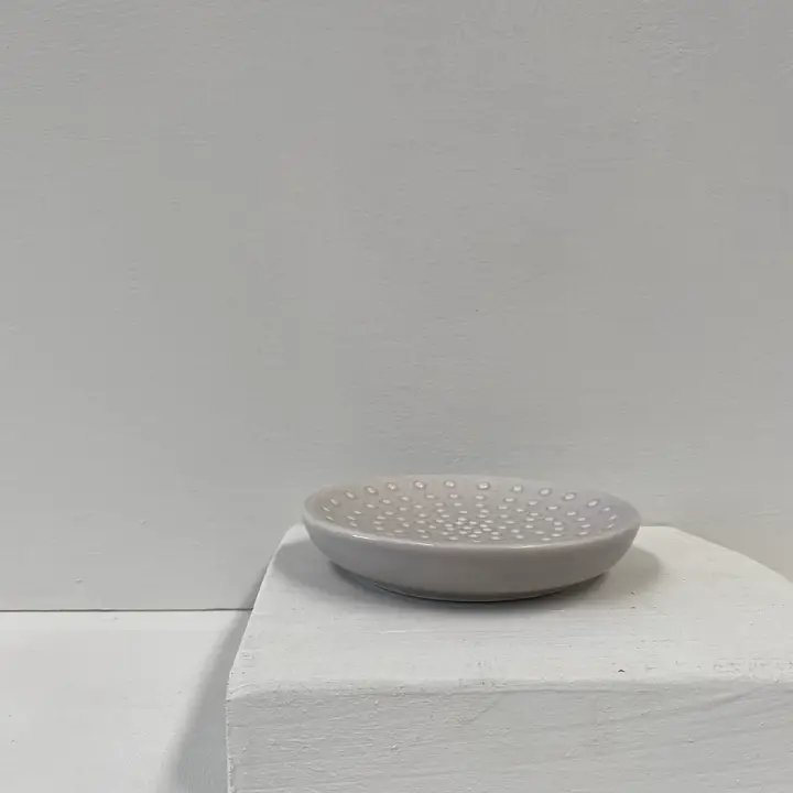 Grå, rund keramikskål med huller i bunden, placeret på en hvid podie mod en grå væg. (2)