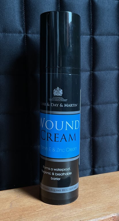 Flaske med sort låg og blå etiket med teksten "Carr & Day & Martin Wound Cream" stående på et lyst bord foran en mørk quiltet baggrund.