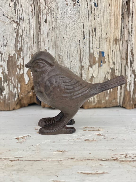 Lille fuglefigur i støbejern placeret på hvidt, slidt træbord med baggrund af afskallet, hvidmalet træpanel.