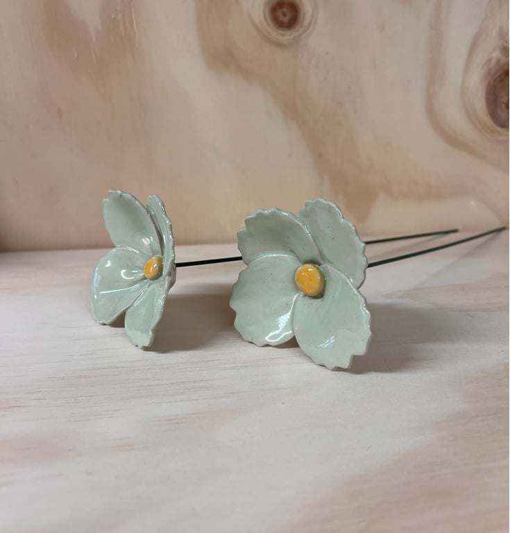 To keramiske blomster med lyseblå blade og gule centre, placeret på træflade med træbaggrund.