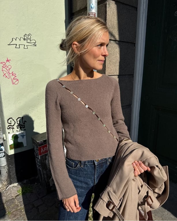 Kvinde iført brun striktrøje og blå jeans, stående udendørs i sollys med beige jakke over armen.