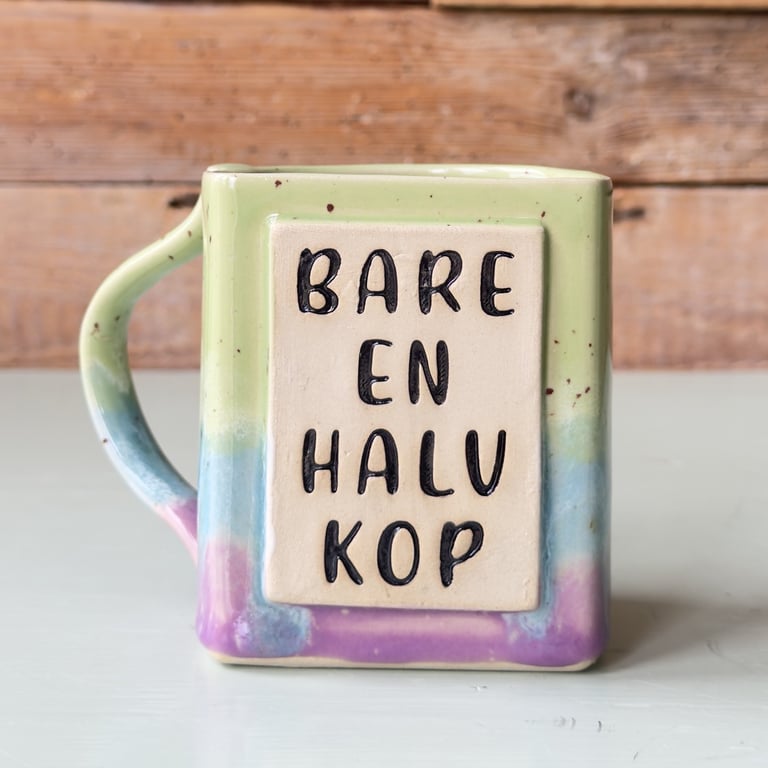 Farverig keramik kop med firkantet form og teksten "BARE EN HALV KOP" på forsiden, placeret på hvid overflade med træbaggrund.