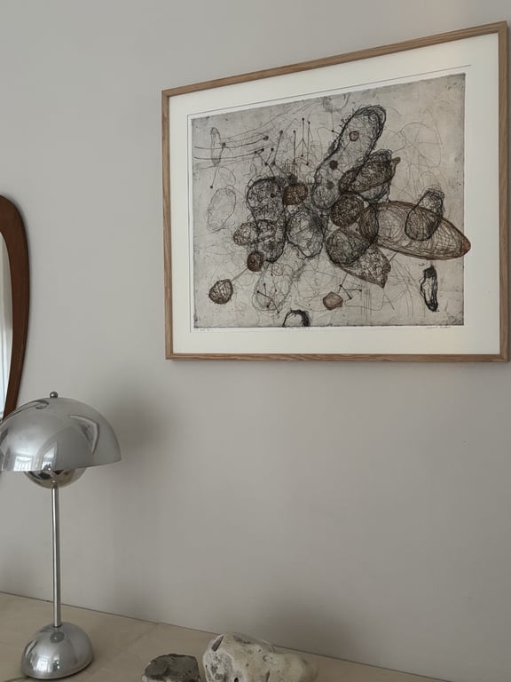 Abstrakt sort-hvid tegning med organiske former i træramme hængende på grå væg over bord med sølvfarvet lampe og sten.