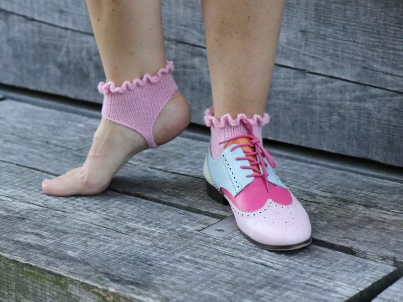 En person iført en farverig sko i rosa, lyseblå og pink på højre fod samt en pink strikket sok med rynkekant, mens venstre fod er bar og også iført en lignende pink strikket sok, stående på et gråt trædæk.