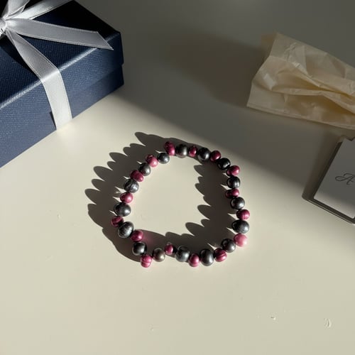 Et armbånd lavet af skinnende pink og sorte perler ligger på et lyst bord, ved siden af en blå gaveæske med hvidt bånd og noget krøllet beige papir. (1)