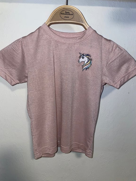 Lyserød kortærmet børnet-shirt med glitrende stof og unicorn-broderi på brystet, hængende på en træbøjle.